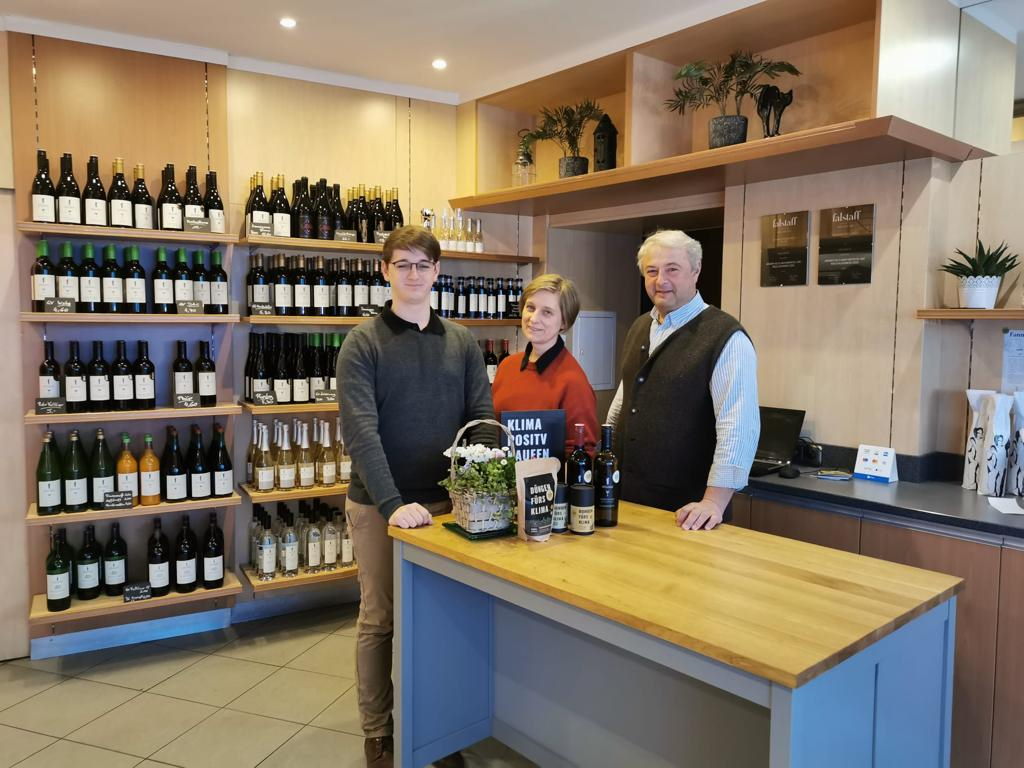 Familie Mößlinger hinter dem Verkaufstisch mit einer Auswahl klimapositiver Produkte. Im Hintergrund die Holzregale des Weingeschäfts Mößlinger gefüllt mit Weinflaschen.